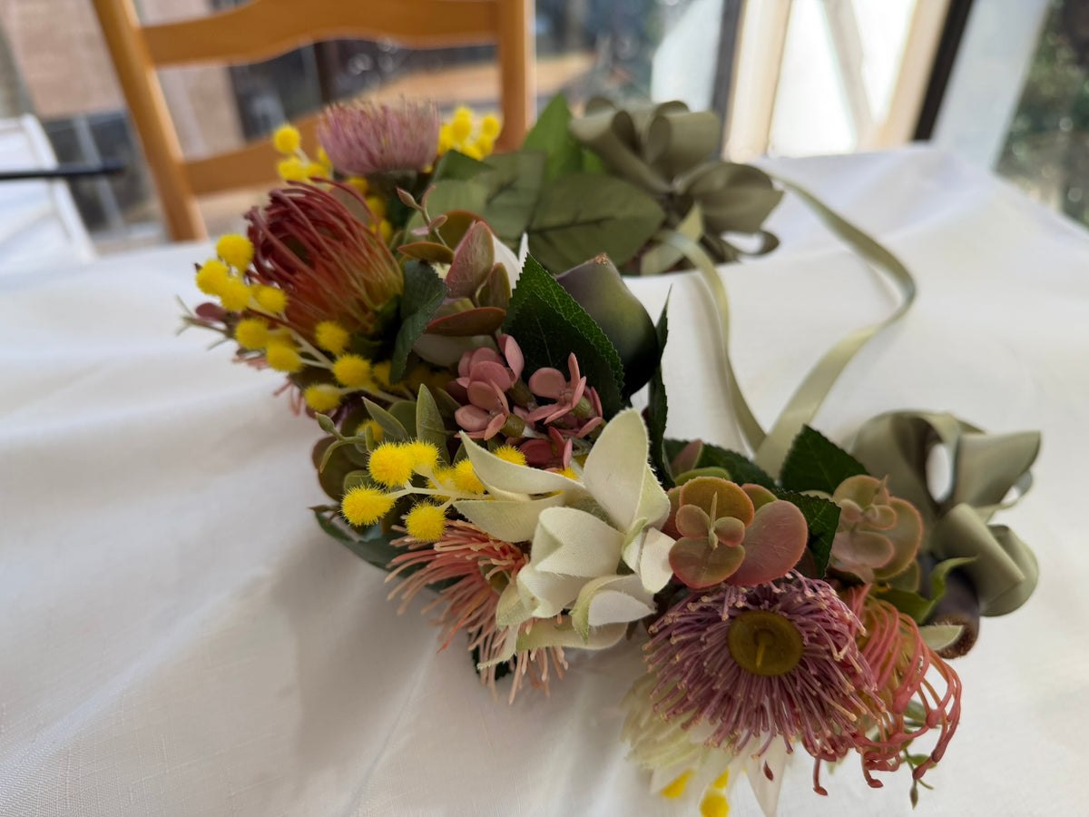 A floral crown with faux protea flowers, green leaves, yellow mimosa, and olive green ribbons, displayed on a white tablecloth.