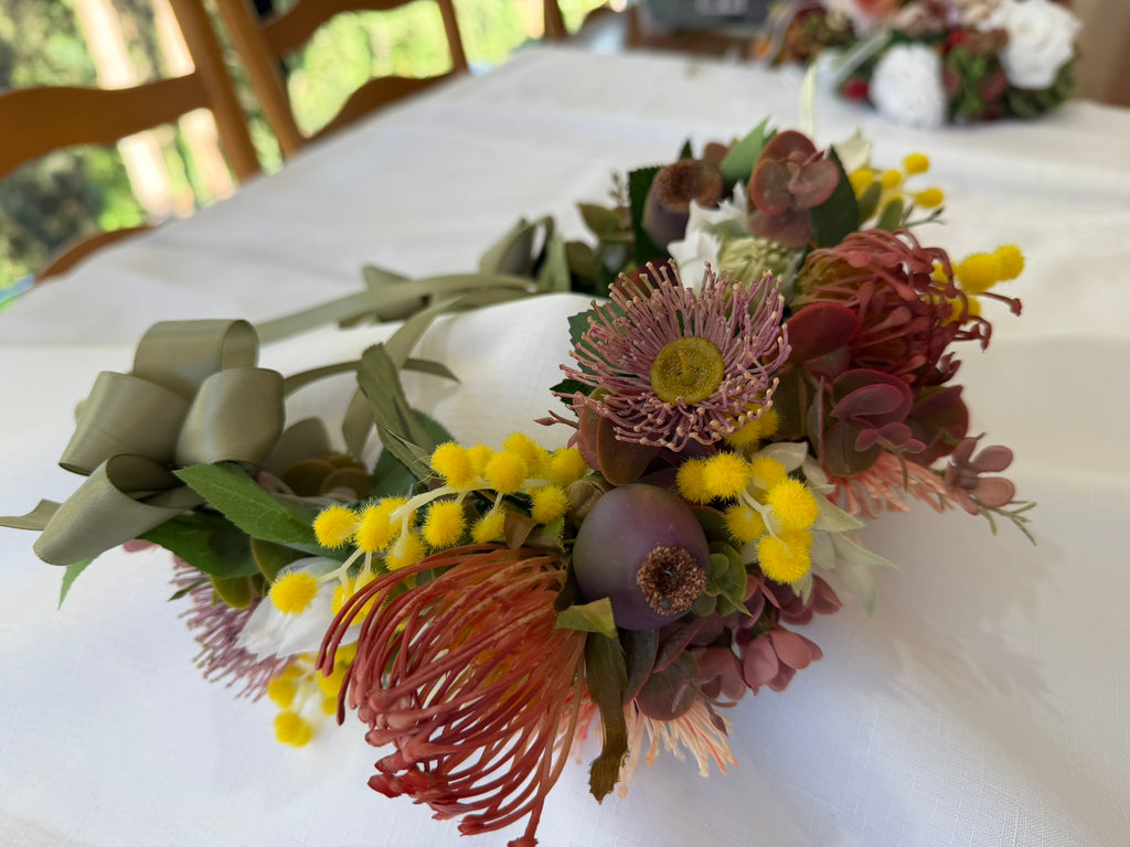 A rustic floral wreath centerpiece featuring yellow mimosa, burgundy protea, green leaves, and sage green ribbon bows, displayed on a white tablecloth.