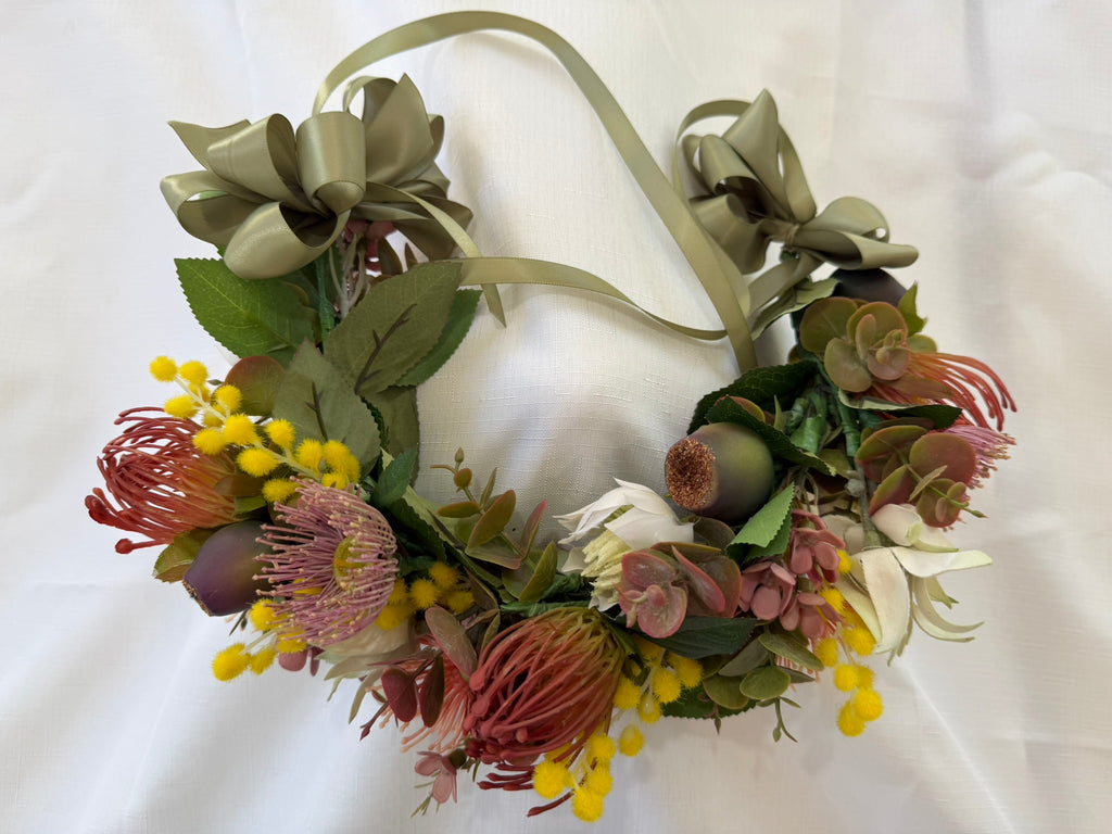 An artificial floral crown wreath featuring olive green satin bows, green leaves, pink and red protea-like flowers, yellow berry clusters, and white lily accents, displayed on a white fabric background.