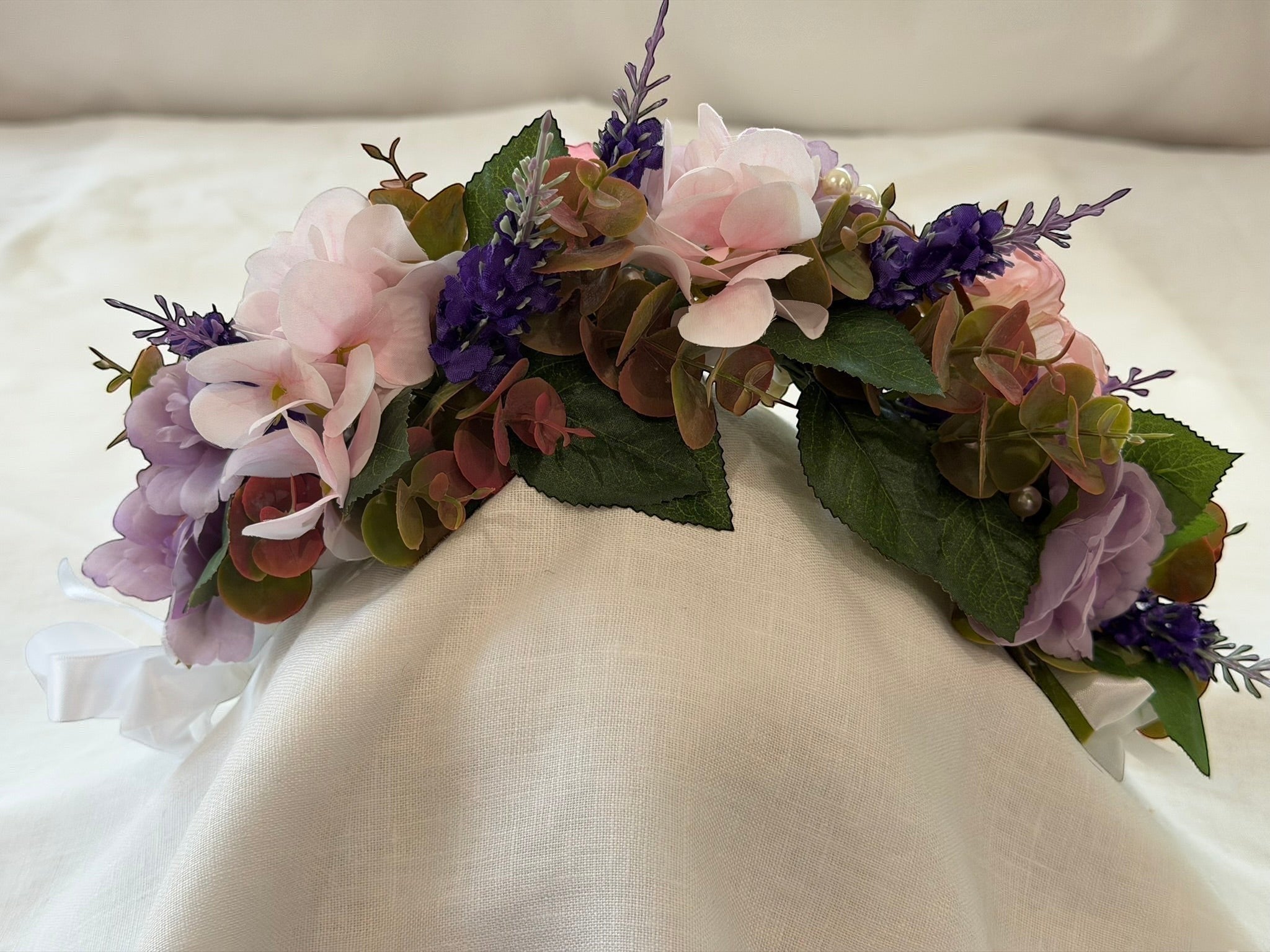 The Velvet Grove floral crown with pink and purple faux flowers, green foliage, and pearls on a white fabric base.