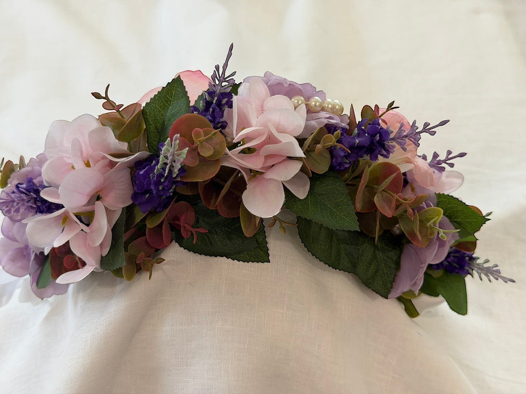 The Velvet Grove floral crown with pink and purple faux flowers, green leaves, and pearl accents on a white fabric.