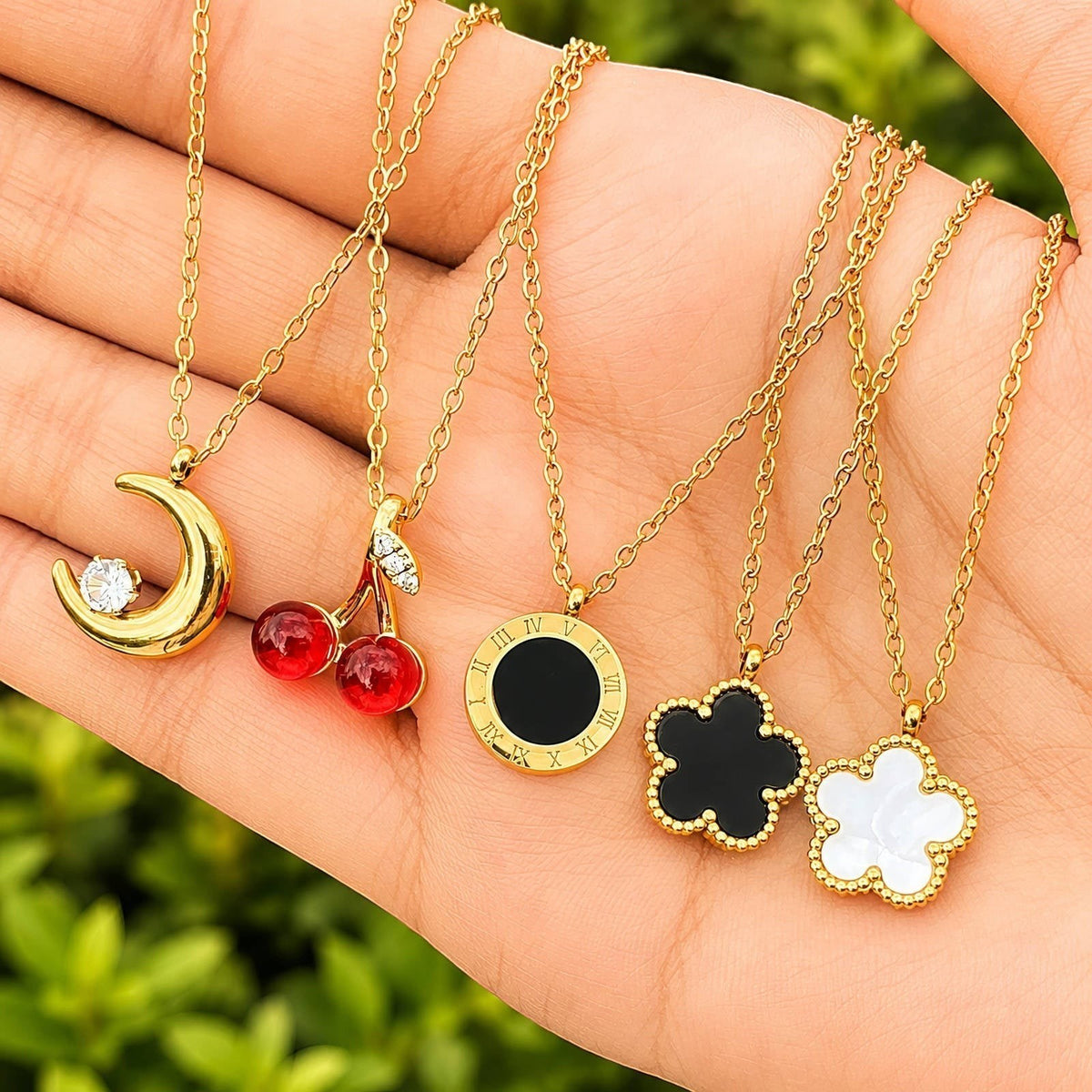 The Velvet Grove gold-tone necklaces featuring cherry, crescent moon, Roman numeral, and flower pendants on a hand.
